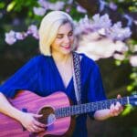 Młoda blond wokalistka gra na gitarze w wiosennym ogrodzie pełnym kwiatów